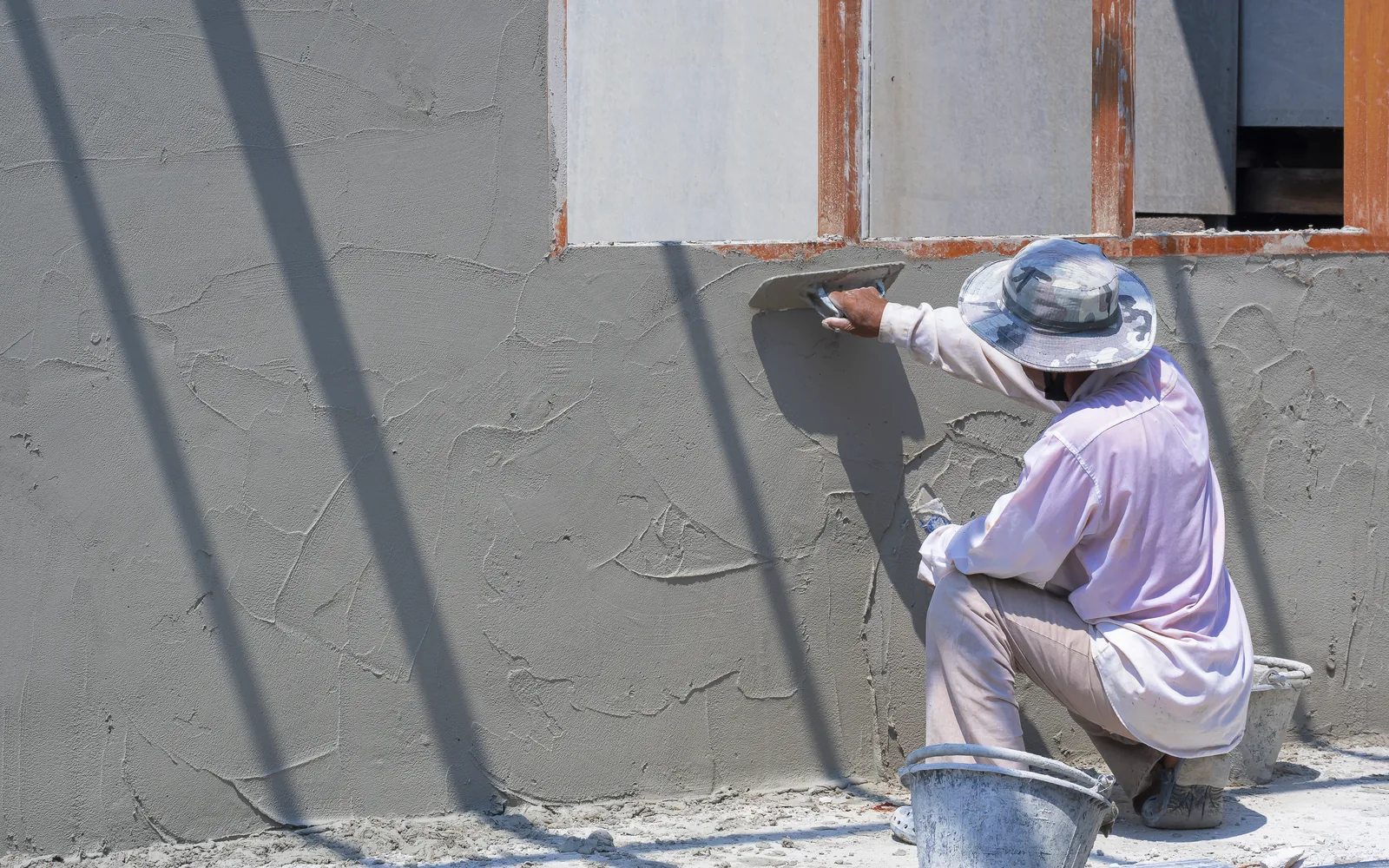 Plasterer in a sun hat troweling a large grey wall